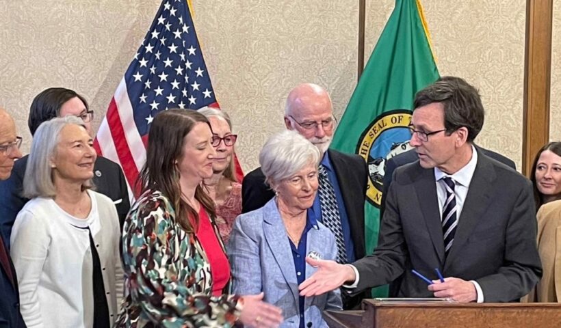 Gov. Bob Ferguson, at podium, goes to shake hands with state Sen. Noel Frame, D-Seattle, at the signing of a bill to make clergy mandatory reporters of child abuse and neglect, on May 2, 2025 in Olympia. Standing between them is Mary Dispenza, a founding member of the Catholic Accountability Project. (Photo by Jerry Cornfield/Washington State Standard)